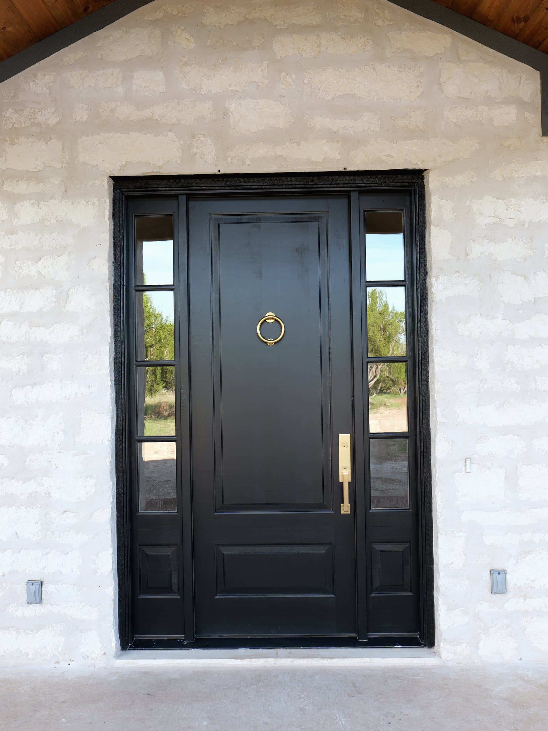 Black front door with a brass knocker beneath a cedar porch gable, set in chopped limestone